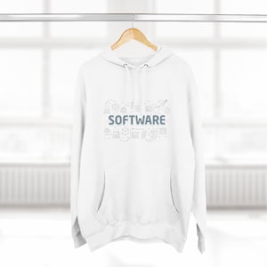 Peut inclure: Sweat-shirt à capuche blanc avec le mot "SOFTWARE" en lettres grises, entouré de petites icônes grises. Le sweat-shirt a une poche avant et une capuche à cordon. Le vêtement est suspendu à un cintre en bois sur un fond clair et flou.