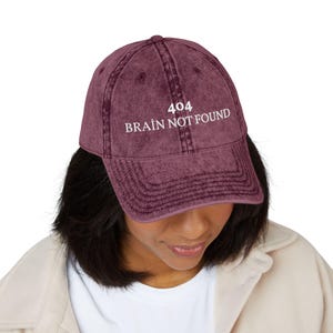 Può includere: Un cappellino da baseball bordeaux con la scritta bianca "404 BRAIN NOT FOUND". Il cappellino ha una visiera curva e un aspetto vintage usurato. Il cappello è indossato da una persona.