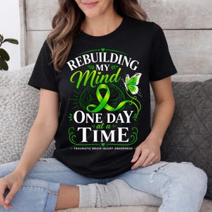 May include: Black t-shirt with the text "Rebuilding My Mind One Day at a Time" in white and green. The design includes a green ribbon, a brain graphic, and a butterfly. The phrase "Traumatic Brain Injury Awareness" is also included.