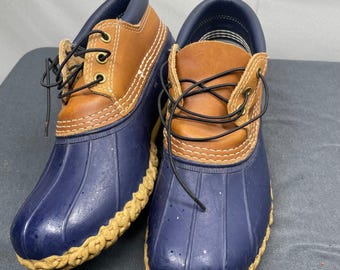 Peut inclure: Une paire de bottes de pluie à hauteur de cheville avec une coque inférieure en caoutchouc bleu foncé à base texturée et une partie supérieure en cuir beige. Les bottes présentent un col rembourré bleu foncé, des œillets en laiton et des lacets noirs.