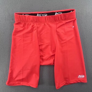 Puede incluir: Pantalones cortos de compresión de color rojo brillante con una cintura elástica negra que presenta el texto blanco ADAMS repetido. Los pantalones tienen un diseño ajustado y liso con un pequeño logotipo ADAMS blanco en la pierna izquierda.