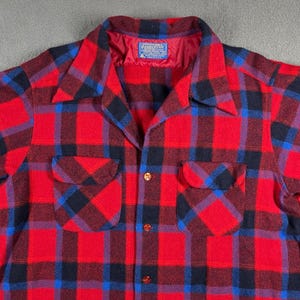 Puede incluir: Una camisa a cuadros roja, negra y azul con dos bolsillos en el pecho. La camisa tiene cuello y botones dorados. La etiqueta dentro del cuello dice "PENDLETON".