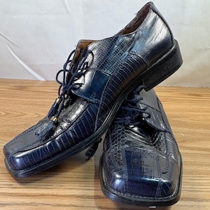 Puede incluir: Un par de zapatos de vestir de cuero azul marino con un patrón texturizado similar al de un cocodrilo. Los zapatos tienen puntera cuadrada, cordones y pequeñas borlas. Los zapatos se exhiben sobre una superficie de madera.