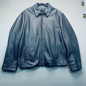 Puede incluir: Chaqueta de cuero negra con cremallera y dos bolsillos delanteros. La chaqueta tiene cuello y está colgada de una percha negra. El cuero parece texturizado.