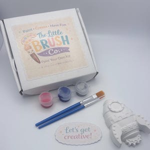 May include: A craft kit from "The Little Brush Co." featuring a white box, paint, brushes, and a rocket-shaped figure. The kit includes paint pots in red, blue, and grey, plus two paintbrushes. A sticker says "Let's get creative!"