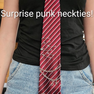May include: A red and white striped necktie featuring silver safety pins, a silver chain with star charms, a spiral metal embellishment, and a soda tab. The text "Surprise punk neckties!" is overlaid on the image. The tie is worn over a black tank top.