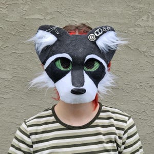 May include: A raccoon-themed mask featuring black and white faux fur, green mesh eye coverings, and a black nose. The mask is decorated with a silver safety pin on the left ear and a metal soda tab with small star charms on the right ear.