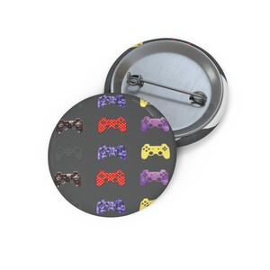 May include: Two round buttons with a pin back. One button features a pattern of colorful game controllers on a dark gray background. The other button is silver with a black rim and a partial view of a colorful design.