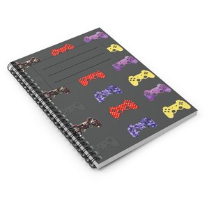 Puede incluir: Un cuaderno de espiral con una cubierta gris oscuro que presenta un patrón de coloridos mandos de videojuegos. Los mandos son de color rojo, morado, amarillo y diseños estampados. El cuaderno tiene una sección rayada para escribir.