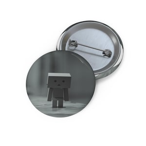 May include: A round button pin featuring a grayscale image of a small cardboard robot figure. The robot has a box-shaped head and a simple body. The pin has a silver metal backing and a safety pin closure.