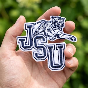Könnte beinhalten: Ein weiß-blaues gesticktes Abzeichen mit den Buchstaben "JSU" und einer Tigerillustration. Der Tiger ist marineblau mit weißen Details. Der Aufnäher wird in einer Hand vor einem verschwommenen grünen Hintergrund gehalten.