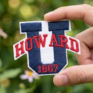 May include: Embroidered patch in the shape of a "U" with the word "HOWARD" in red letters. The "U" is navy blue with a white center and the number "1867" in red. The patch is held against a blurred green background.