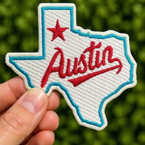 May include: Embroidered patch in the shape of Texas, with a white background and a blue border. The word "Austin" is stitched in red script across the centre, and a red star is in the upper left corner. The patch is held against a blurred green background.