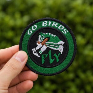 May include: A round, embroidered patch with a green and black design. The patch features the words "GO BIRDS" in white, arched around the top. A football player in green and white is in the center, with the word "FLY" in green below.