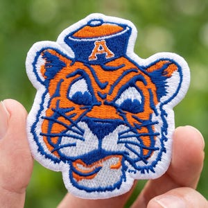 Könnte beinhalten: Gestickter Aufnäher mit einem Tiger-Maskottchen-Kopf in Orange, Blau und Weiß. Der Tiger trägt einen blauen Hut mit orangefarbenem Rand und dem Buchstaben "A". Der Aufnäher hat einen weißen Rand und wird vor einem verschwommenen grünen Hintergrund gehalten.