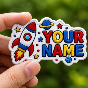Puede incluir: Un parche bordado colorido con un cohete, estrellas y un planeta. El parche muestra las palabras "YOUR NAME" en letras brillantes. El diseño está sobre un fondo blanco.