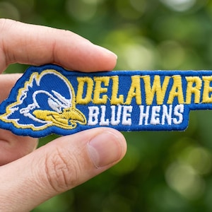 May include: Embroidered patch featuring the Delaware Blue Hens logo. The patch is rectangular with a blue background and yellow and white lettering that reads "DELAWARE BLUE HENS". The logo includes a blue and white bird with a yellow beak.