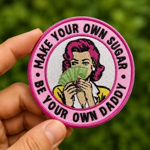 May include: A circular, pink embroidered patch with the text "MAKE YOUR OWN SUGAR BE YOUR OWN DADDY." The patch features a retro illustration of a woman holding money. The woman has pink hair and is wearing a yellow top.