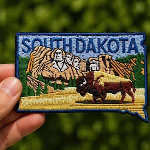 May include: Embroidered patch featuring the words "SOUTH DAKOTA" in blue. The patch depicts Mount Rushmore and a bison on a field of yellow. The patch has a blue border and is shaped like the state of South Dakota.