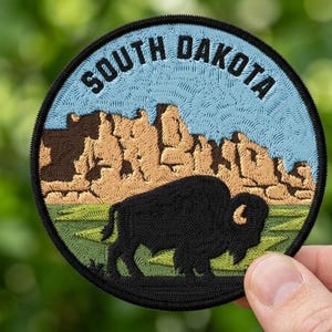 May include: A circular embroidered patch featuring a black bison standing in a green field against a backdrop of tan rock formations and a light blue sky. The text SOUTH DAKOTA is stitched in black capital letters along the top edge of the patch.