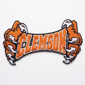 May include: An embroidered patch featuring the word CLEMSON in orange block letters with a black outline. The text is flanked on both sides by large, detailed orange and white tiger paws with sharp, curved white claws, all bordered in black.