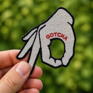 Puede incluir: Parche bordado con la forma de una mano haciendo el gesto de 'OK', con la palabra 'GOTCHA' en letras rojas. El parche tiene un fondo gris con contorno negro.