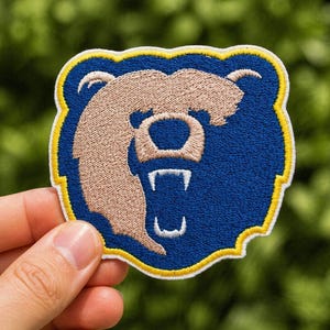 May include: Embroidered patch featuring a bear head design. The patch has a blue background with a tan bear head and white teeth. The patch is outlined in yellow and white.