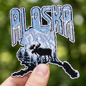 Könnte beinhalten: Gestickter Alaska-Aufnäher. Der Aufnäher hat die Form des Bundesstaates und zeigt den Namen „Alaska“ in einer stilisierten Schriftart. Er zeigt eine Bergkette, einen Elch und Bäume in Blau, Weiß und Schwarz mit einem schwarzen Rand.