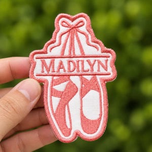 Puede incluir: Parche bordado rosa coral con zapatillas de ballet, con el nombre "MADILYN" en una pancarta. El parche tiene detalles en blanco y un borde blanco. El parche está sujeto por una mano sobre un fondo verde borroso.