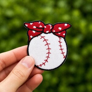 May include: Embroidered patch of a baseball design with a red and white polka dot bow. The baseball is white with red stitching, and the patch has a black border. A fun accessory for sports fans.