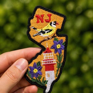 Könnte beinhalten: Gestickter Aufnäher in Form von New Jersey mit einem gelben Vogel, lila Blumen und einem Leuchtturm. Der Aufnäher hat oben die Buchstaben "NJ" in Rot. Der Aufnäher wird in einer Hand vor einem verschwommenen grünen Hintergrund gehalten.