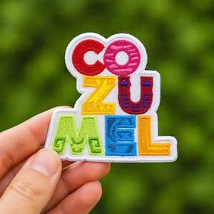 Op de afbeelding: Geborduurde patch met het woord "COZUMEL" in kleurrijke blokletters. De patch heeft een witte rand en een groene achtergrond. De letters zijn rood, roze, geel, blauw en oranje.