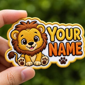 May include: Embroidered patch featuring a cartoon lion with a brown mane and paws. The patch has the text "YOUR NAME" in yellow and orange. A paw print is also included. The patch is held in a hand.