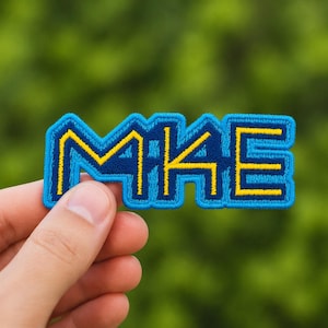 Puede incluir: Parche bordado con las letras "MKE" en una fuente audaz y estilizada. El parche presenta un fondo azul con costuras amarillas y azules, y un borde azul claro. El parche se sostiene en una mano.