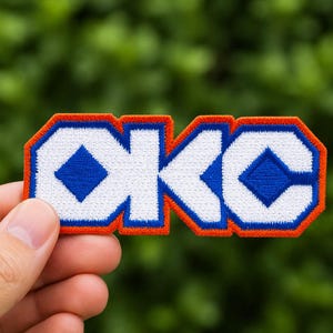 May include: Embroidered patch with the letters "OKC" in white, outlined in blue and orange. The patch features a diamond shape within the "O" and "C". The patch is held against a blurred green background.