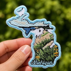 May include: Embroidered patch in the shape of the state of Michigan. The patch features a light blue border and depicts the state's landscape with green, blue, and white thread. The word "Michigan" is embroidered in cursive.