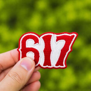 Puede incluir: Un parche bordado rojo y blanco con el número "617". El parche tiene un contorno blanco y se sostiene sobre un fondo verde borroso. El parche es un diseño estilizado.