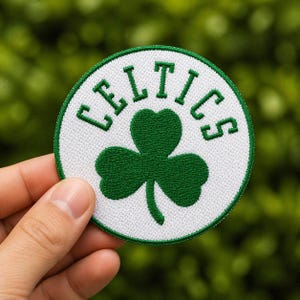 May include: A round patch featuring the Boston Celtics logo. The patch has a white background with a green shamrock in the center. The team name, "CELTICS", is embroidered in green around the shamrock.