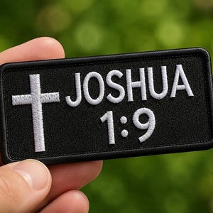 May include: Black rectangular patch with white embroidered text and a cross. The text reads "JOSHUA 1:9". The patch has a black border and is held against a blurred green background.
