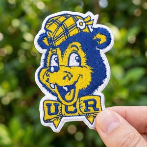 May include: Embroidered patch featuring a cartoon bear wearing a plaid hat. The bear is yellow and blue with a smiling face. The patch has the letters "UCR" in a banner at the bottom. The patch is held in a hand.