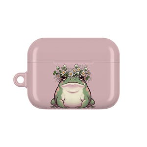 Puede incluir: Una funda AirPod rosa claro con un dibujo de una rana con una corona de flores de margarita. La rana es verde y blanca con ojos negros. La funda tiene un lazo para sujetar.