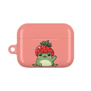 May include: A coral-colored AirPod case featuring a cartoon frog wearing a strawberry hat. The frog is green with a white belly, black eyes, and a small smile. The case has a smooth finish and a small loop for attaching a keychain.