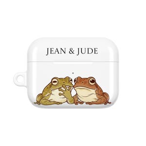 Op de afbeelding: Witte AirPod-hoes met een cartoon illustratie van twee kikkers. Eén kikker is groen, de andere bruin. De tekst "JEAN & JUDE" staat in het zwart bovenaan. De hoes heeft een kleine lus aan de zijkant.