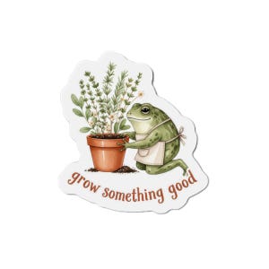 Pode incluir: Um adesivo recortado com uma rã verde usando um avental bege, ajoelhada ao lado de um vaso de terracota cheio de ervas verdes e pequenas flores brancas. O texto "grow something good" está impresso em letras castanhas abaixo.