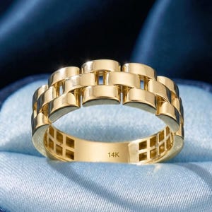Puede incluir: Un anillo dorado con un diseño de eslabones rectangulares, sobre un fondo azul. El anillo está marcado con "14K". El anillo se muestra en una caja de joyería azul.