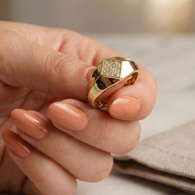 May include: A gold ring with a square face set with small, clear stones. The ring has a geometric design with a wide band. The ring is held in a hand with peach-coloured nail polish.