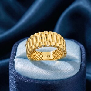 May include: A gold-colored ring with a textured, repeating rectangular design. The ring is marked with "14K" on the inside. The ring is displayed on a blue satin surface.