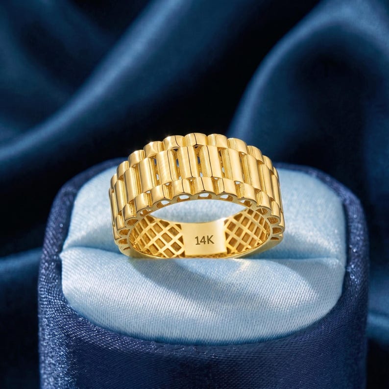 May include: A gold-colored ring with a textured, repeating rectangular design. The ring is marked with "14K" on the inside. The ring is displayed on a blue satin surface.