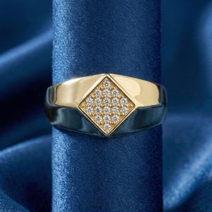 May include: A gold and silver ring featuring a diamond-shaped gold centre set with small, clear gemstones. The ring's band is silver, and it is displayed against a backdrop of blue satin fabric. The ring is a piece of jewellery.