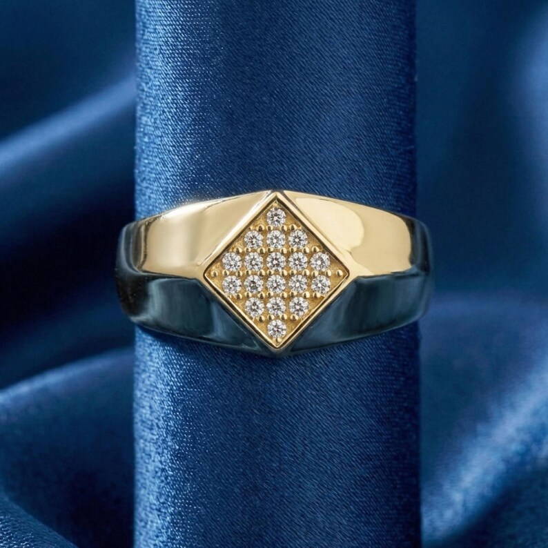 May include: A gold and silver ring featuring a diamond-shaped gold centre set with small, clear gemstones. The ring's band is silver, and it is displayed against a backdrop of blue satin fabric. The ring is a piece of jewellery.